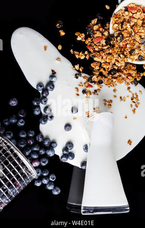 Joghurt aus Glas Krug verschüttet, Blaubeeren gegossen aus Stahl stainer und Müsli gegossen und aus Emaille Schüssel verstreut gemischt zusammen in ein Chaos auf Stockfoto