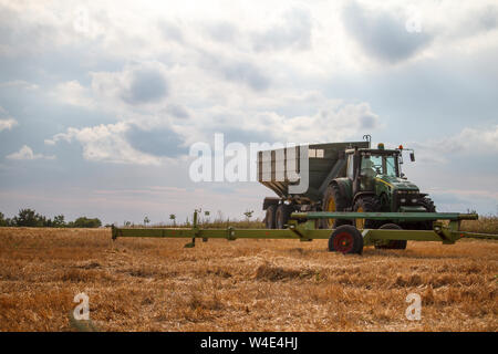 Charkow, Ukraine - Juli 16, 2019: ein Traktor für das Sammeln von Weizen von einem Mähdrescher auf einem Feld von Gemähten Weizen gegen einen blauen bewölkten Himmel steht Stockfoto