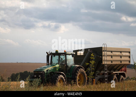 Charkow, Ukraine - Juli 16, 2019: ein Traktor für das Sammeln von Weizen von einem Mähdrescher auf einem Feld von Gemähten Weizen gegen einen blauen bewölkten Himmel steht Stockfoto