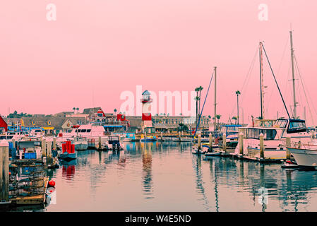 Blue sea im Hafen mit roten und weißen Leuchtturm und angedockten Boote unter eine blass rosa Himmel. Stockfoto