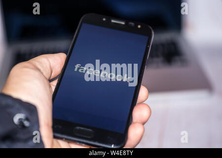 Nizhyn, Ukraine/Juli -16-2019: Junger Mann halten-Smartphone in der Hand und Facebook App mit Laptop auf Hintergrund verwenden. Social Media und Körperschaftlichen conce Stockfoto