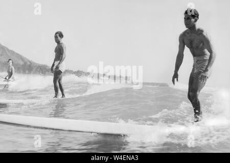 Surfer auf hölzernen Longboards und eine Frau in einem Outrigger Kanu Reiten am Strand von Waikiki in Honolulu, Hawaii, im Frühjahr 1936. Stockfoto