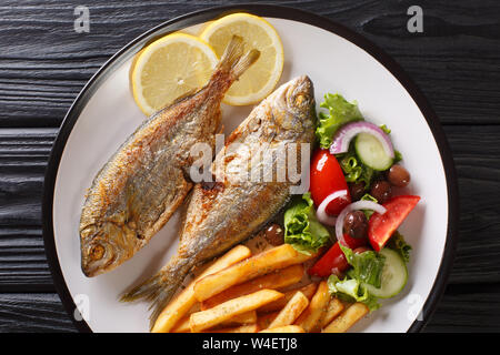 Teil der Fried Salema porgy Fisch mit Zitrone, Pommes frites und frisches Gemüse Salat close-up auf einem Teller auf den Tisch. Horizontal oben Ansicht von oben Stockfoto