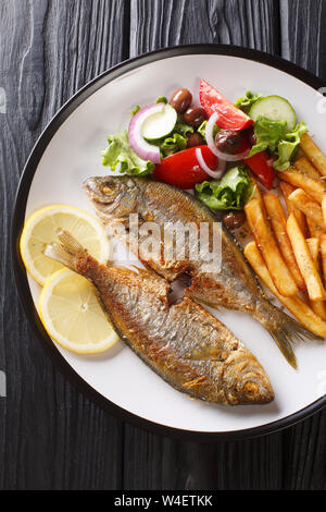 Teil der Fried Salema porgy Fisch mit Zitrone, Pommes frites und frisches Gemüse Salat close-up auf einem Teller auf den Tisch. Vertikal oben Ansicht von oben Stockfoto