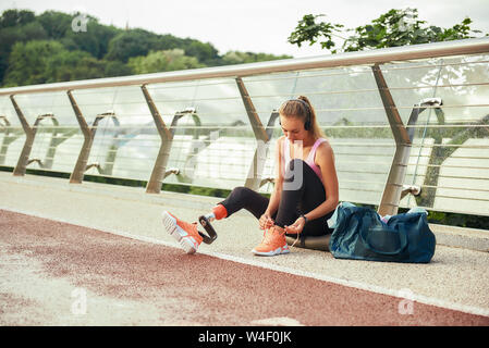 Immer bereit zu laufen. Junge Frau in Sportkleidung und Kopfhörer mit beinprothese Band ihre Schnürsenkel beim Sitzen auf der Brücke. Behinderten Sport Konzept. Motivation. Gesunder Lebensstil Stockfoto