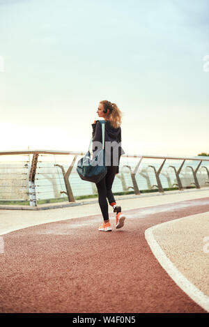 Morgen Workout. Zurück Blick auf junge behinderte Frau im Sport Kleidung mit beinprothese hält ihre Sporttasche, während auf der Brücke stehen am Morgen. Behinderten Sport Konzept. Motivation. Gesunder Lebensstil Stockfoto