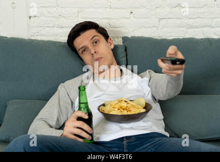 Lifestyle Portrait der junge Mann gelangweilt auf der Couch mit Fernbedienung zapping für Film oder Sport live. Suchen desinteressiert Bier trinkt. Bewegungsmangel und Stockfoto