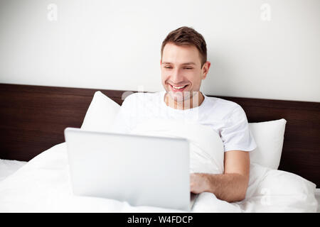 Glückliche junge Mann im Bett sitzend mit seinem Computer Stockfoto
