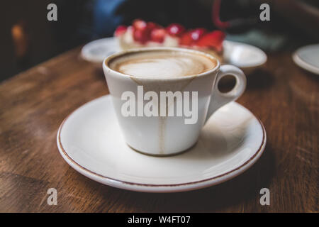 Weiße Tasse Latte Kaffee mit Kuchen mit Erdbeeren auf einer hölzernen vintage Tisch in einem netten Café. Touristische Sehenswürdigkeiten Stockfoto