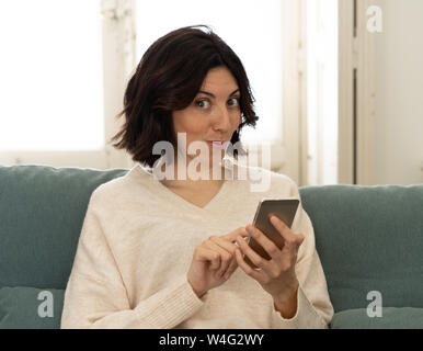 Lifestyle Portrait von attraktiven Frau auf Smart Handy Nachrichten senden, chatten oder in sozialen Medien zufrieden lächelnd auf der Couch zu Hause. In Leis Stockfoto