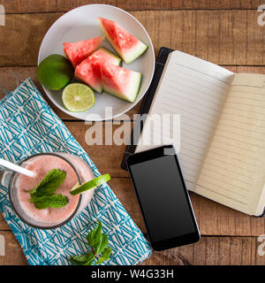 Ein flach (Quadrat) Frühstück Tischplatte Foto mit Wassermelone smoothie Telefon, Buch- und geschnittenes Obst auf dem Teller - Nahrung und Diät Konzept Bild. Stockfoto