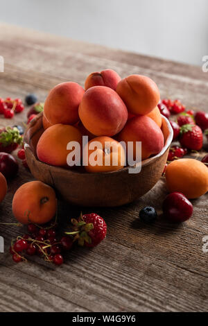 Reifen köstliche saisonale Beeren um Schale mit frischen Aprikosen auf Holztisch isoliert auf Grau verstreut Stockfoto