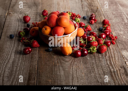 Reifen köstliche saisonale Beeren um Schüssel mit Aprikosen auf hölzernen Tisch verstreut Stockfoto