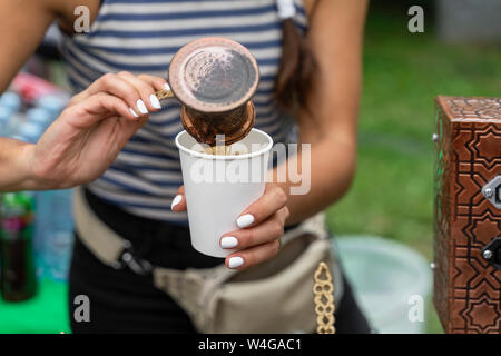 Hände von Mädchen gießt Türkische aromatischen Kaffee mit Schaum in das verfügbare Papier Cup. Vorbereitung kochen in Kupfer Turk auf heißem Sand, arabischer Kaffee Stockfoto