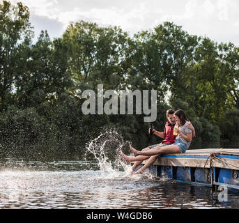 Junges Paar mit einem Drink und Spritzen mit Wasser am Steg an einem entfernten See Stockfoto