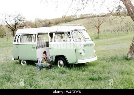 Junge Frau Studium Karte, im Gras sitzen, vor ihr Wohnmobil Stockfoto