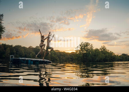 Junge Paare springen von badeplattform auf einem See, halten sich an den Händen Stockfoto