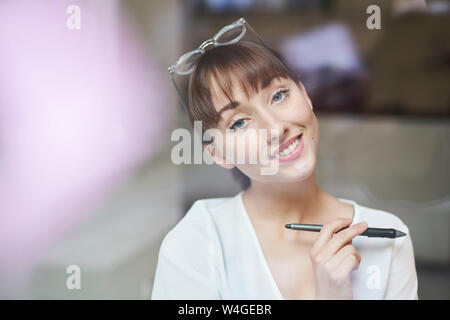 Portrait von lächelnden jungen Geschäftsfrau mit Brille und Bleistift hinter Glasscheibe Stockfoto