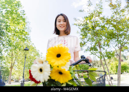 Portrait von lächelnden jungen Frau mit Blumen und Fahrrad im Park Stockfoto