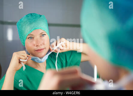 Frau in scrubs Suchen in Spiegel setzen auf Maske Stockfoto