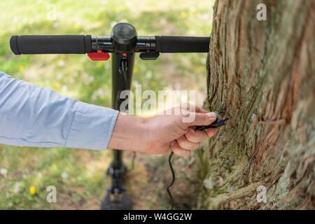 Man' Hand, die Stecker des E-Scooter vor Baumstamm, close-up Stockfoto