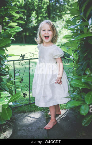 Portrait von glücklichen kleinen Mädchen mit weißem Kleid steht in der Nähe von Garden Gate Stockfoto