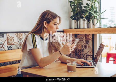 Junge Frau mit dem Handy und dem Laptop im Cafe Stockfoto