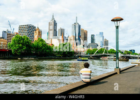 Mann sitzt am Yarra River in Richtung Wolkenkratzer in Melbourne, Victoria, Australien suchen Stockfoto