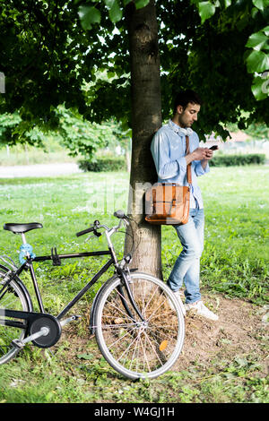 Junger Mann mit Smartphone Fahrrad, Kopfhörer um den Hals, stehend unter Baum Stockfoto