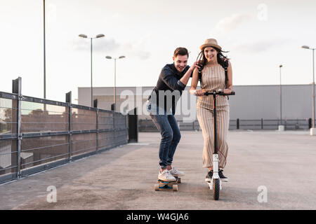 Junger Mann und eine Frau reiten auf Longboard und Elektroroller auf Parkdeck Stockfoto