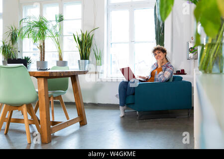 Lächelnde Frau mit Laptop im Sessel im Büro Stockfoto