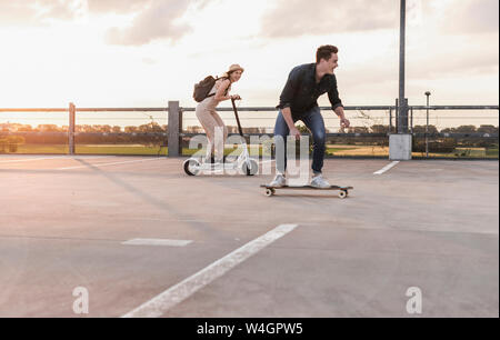 Junger Mann und eine Frau reiten auf Longboard und Elektroroller auf Parkdeck bei Sonnenuntergang Stockfoto