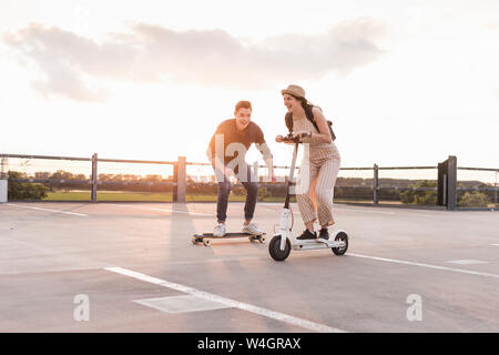 Junger Mann und eine Frau reiten auf Longboard und Elektroroller auf Parkdeck bei Sonnenuntergang Stockfoto