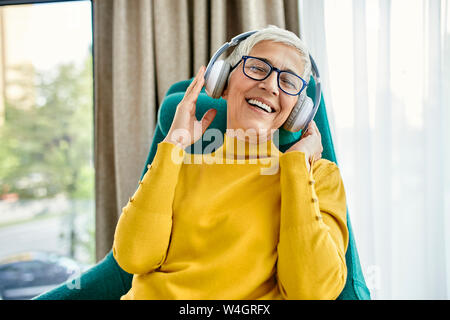 Ältere Frau im Sessel sitzen, Musik hören mit Kopfhörern Stockfoto