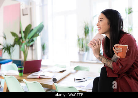 Lächelnde Frau mit Handy sitzt am Tisch in modernen Büro Stockfoto