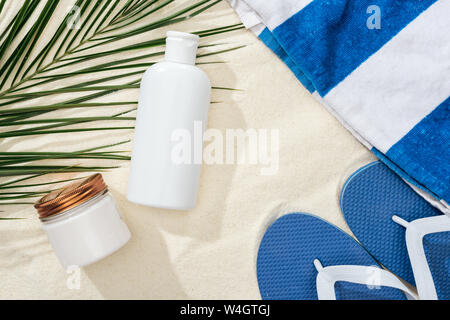 Weiß sunscreen Lotion und Creme in der Nähe von Green Palm Leaf auf Sand mit blauen Flip Flops und Handtuch Stockfoto