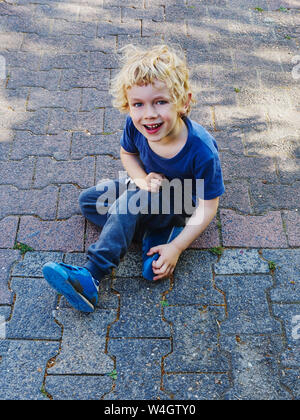 Portrait von lächelnden kleinen Jungen in Blau gekleidet sitzen auf Pflaster Stockfoto