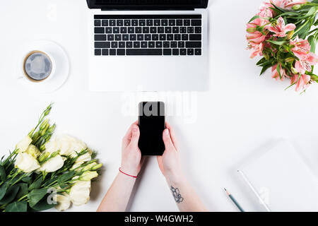 7/8-Ansicht von Frau mit Smartphone mit leerer Bildschirm in der Nähe von Laptop, Tasse Kaffee und Blumensträuße auf Weiß Stockfoto