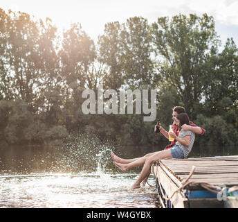 Glückliches junges Paar mit einem Drink und Spritzen mit Wasser am Steg an einem entfernten See Stockfoto