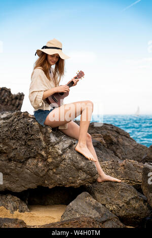 Lächelnden jungen Frau Sommer Hut sitzen auf Rock spielen Ukulele Stockfoto