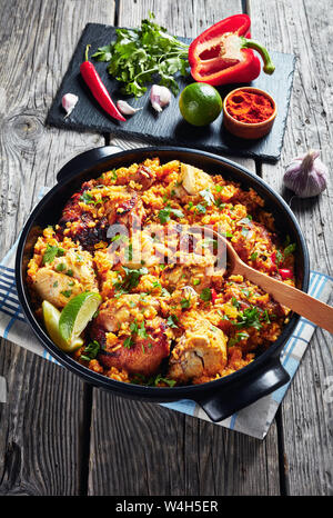 Arroz con Pollo, spanische Küche, Reis mit Huhn und Gemüse in einem schwarzen Topf auf einem hölzernen rustikalen Tisch mit Zutaten auf einer Schiefertafel, Verti Stockfoto