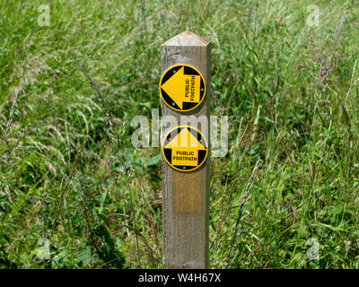 Öffentlichen Fußweg Zeichen mit zwei gelben Pfeile auf dem kleinen schwarzen Scheiben auf einem kurzen hölzernen Pfosten gegen eine lange Gras Hintergrund. Eins vor, links Stockfoto