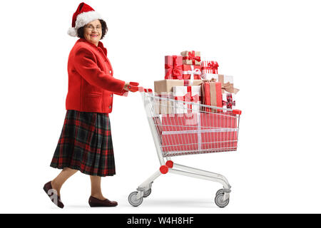 Volle Länge geschossen von einer älteren Frau mit einer Mütze treibt ein Warenkorb mit Geschenken und mit Blick auf die Kamera auf weißem Hintergrund Stockfoto