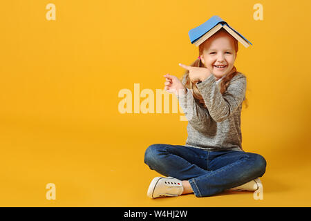 Freundliche attraktive kleine Schüler Mädchen sitzt auf dem Boden mit einem Buch auf den Kopf und zeigt auf die Seite. Der Begriff der Bildung und Schule. Stockfoto