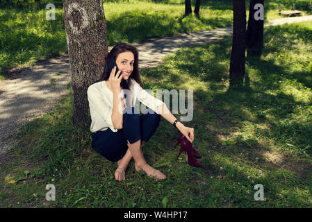 Business Frau sitzt unter dem Baum lächelnd Holding ihre Schuhe in die Hand und sprechen über das Telefon in einem öffentlichen Park selektiven Fokus Stockfoto