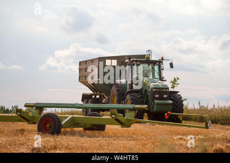 Charkow, Ukraine - Juli 16, 2019: ein Traktor für das Sammeln von Weizen von einem Mähdrescher auf einem Feld von Gemähten Weizen gegen einen blauen bewölkten Himmel steht Stockfoto