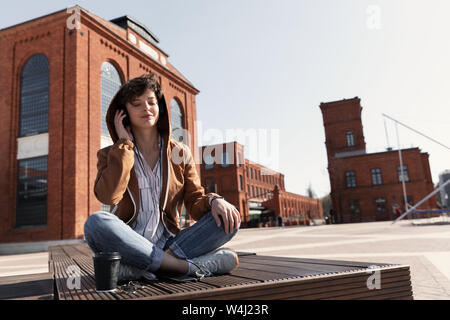 Ein junges Mädchen mit einem stilvollen Kurzhaarschnitt sitzt auf einer Bank und hört Musik über Kopfhörer. Stockfoto