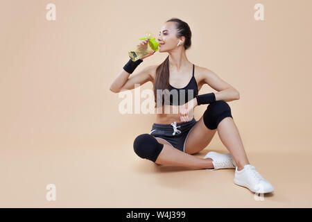 Ein niedliches athletische brunette Mädchen, in einer enganliegenden Sport einheitliche, sitzt, liegt nach dem Training, in schwarz Kniescheiben, mit einer Flasche für Sporternährung. Stockfoto