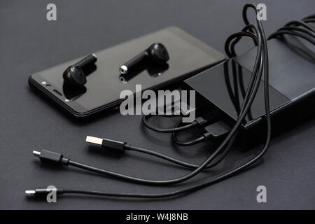 Es ist ein Smartphone, ein Headset und eine Bank mit Ladekabel auf einem dunklen Hintergrund. Stockfoto