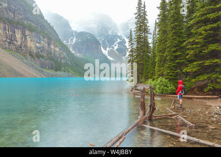 Mann in roter Regenjacke steht durch den Moraine Lake Shore unter dem Regen an einem Sommermorgen, Banff National Park, Alberta, Kanada. Stockfoto